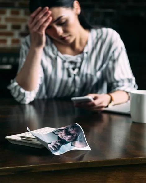 Concerned woman sitting at a table with a phone, holding her head in distress, contemplating calling an addiction intervention helpline for help with a loved one’s struggle.